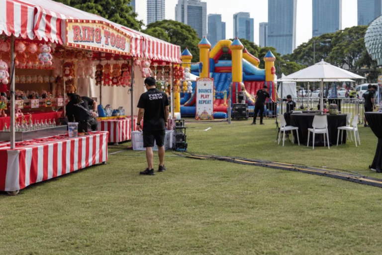 Event staff prepare game stalls, a bouncy castle, tables, and equipment for a carnival rental in Singapore at an outdoor venue